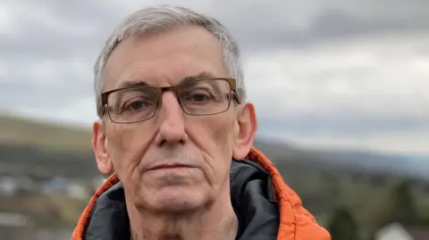 Chris Austin stands in front of the Ffos-y -Fran mine in Merthyr Tydfil. It is a head shot, he has short grey hair and glasses on. 