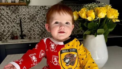 Baby Beatrice sits atop a kitchen counter wearing a half red and half yellow baby gro. Each side has the respective badges on. She has short red hair and is smiling slightly at the camera, with her hands outstretched. There is a vase of yellow flowers behind her.