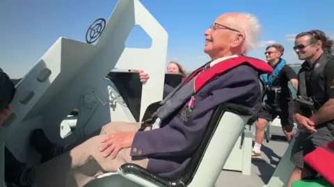 Ken has a seat on the front of the boat as it speeds along the water, a big smile on his face. He has white hair and glasses and wears his uniform, medals, and a  life jacket.