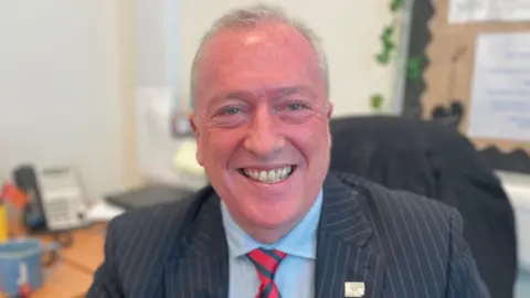A man with short gray hair in a dark red and green tie and a dark brown suit jacket. He smiled and sat at his desk in the office with a pinboard behind him.