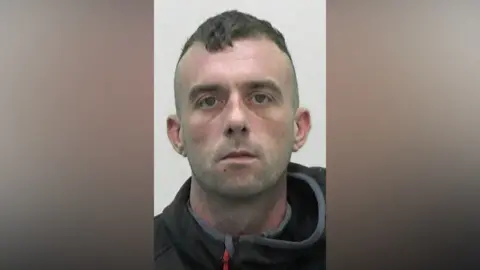 Mugshot of Toward. He has faint stubble and short dark hair on top in a drooped quiff with the sides shaved. He has a vacant look in his eyes.