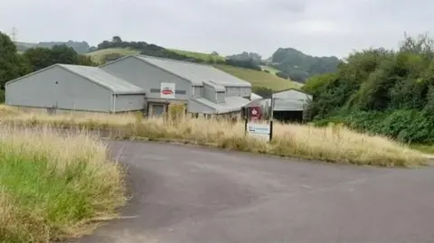 Google An industrial building in a rural setting. The large grey building is surrounded by dry grass and there is a road leading to it. 