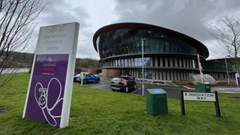 The headquarters of Caudwell Children, a circular glass-fronted building with a car park in front of it. There is a totem on a grass verge which has a white butterfly emblem on a purple background and a white sign with black writing that reads Innovation Way.