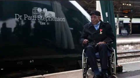 Dr Stephenson in a wheelchair being pictured next to a train with his name on it. He is wearing a black jacket with a gold medal on his lapel. He is also wearing a visor seen during the covid pandemic. 