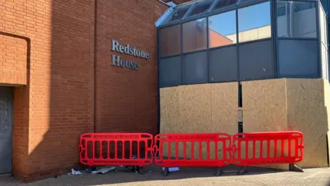 A brick-built block of flats with several storeys, with a glass entrance porch and "Redstone House" in silver lettering on a brick wall at right angles to the main building.  There is wooden boarding around the entrance porch, and a low red temporary fence in front of the entrance.