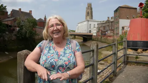 Karl Bird / BBC Jane Keightly stood on the side of a street next to a canal. She is smiling at the camera, leaning against a railing. She has shoulder length blonde hair and is wearing glasses, a blue, green and red, floral-patterned top and a rucksack on her back. Buildings can be seen behind her lining the canal. The sky is blue.