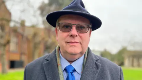 Emma Baugh/BBC Paul Van der Hulke who is wearing a Trilby-style hat and a grey overcoat over a shirt and tie. He is wearing glasses. He is standing outside with grass and buildings behind him. 