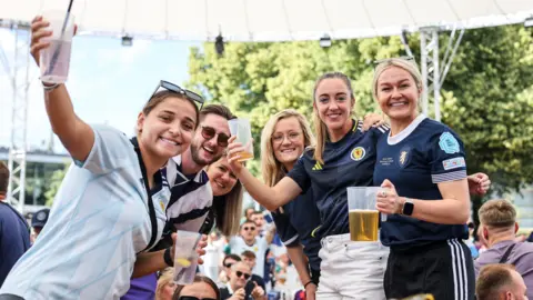 Getty Images Scotland fans at Olympiapark in Munich during the Euro 2024 match with Germany. Five women and one man are standing up, holding drinks and smiling at the camera. They are all wearing Scotland tops.