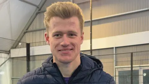 Ruari is wearing a navy puffer jacket. He has short blonde hair. Behind him is the fence of a Padel court. The court is inside a large tin like shed. 