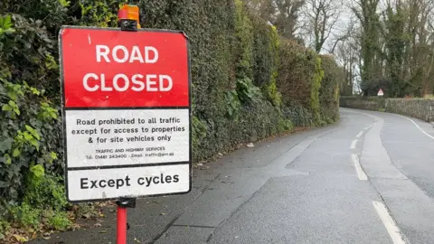 A road closed sign on a road in Guernsey. The sign is red and white. It says the road is closed for road users except for cyclists. One side of the road has hedging along it while the other has a stone wall running along it.