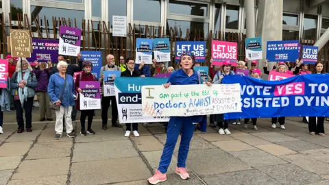 Demonstrators outside the parliament building. A large group of pro-assisted dying demonstrators are in the background and a woman with a banner saying she is a doctor opposed to the legislation is standing in front of them