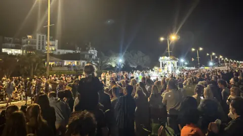 Submitted The crowds lining the road are five deep. Spectators are watching a float to the right of the picture. It is dark but there are street lamps.