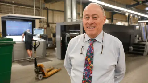 A man in a shirt and floral tie stands in front of printing machinery