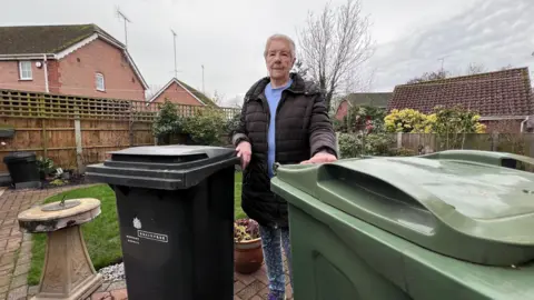 A lady in her seventies standing in a garden looking at the camera with two large wheelie bins next to her.