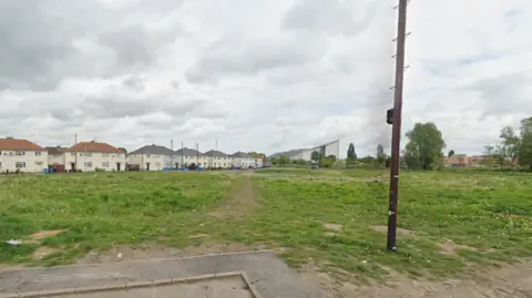 Google A piece of disused grassland. White houses can be seen in the distance. The sky is overcast. 