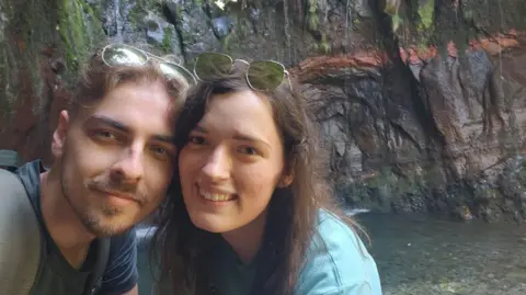 Liam Watson A man with short brown hair and a beard stands next to woman with long brown curly hair and brown eyes. They both have sunglasses their heads and are smiling at the camera for a selfie. Behind them are dark rock cliffs and a river below.