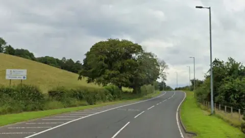 A71 east of Newmilns. The road curves round a corner in the distance and has the word "SLOW" on the road. Trees and bushes line the road and a field is seen on one side. 