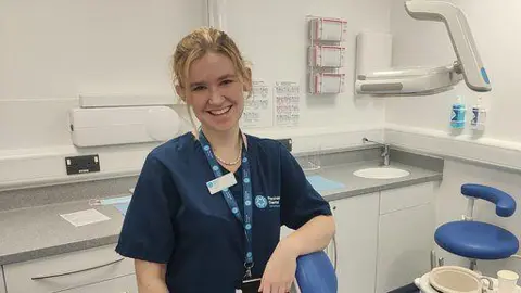 University of Plymouth Ellie has her hair tied back and is wearing a blue top with a lanyard which is also blue. She has her arm over a blue chair and is smiling. She is in a dental room.