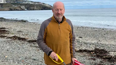 Bill Dale, a man with a grey beard, wears a brown and grey fleece and smiles. He is on the beach and you can see the headland in the background.