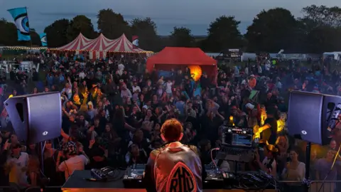 A small community festival scene at dusk. The illuminated back of a DJ can be seen as he performs on a stage. There is a crowd in front of him. Red and white circus tents and other canopies can be seen dotted among the crowd of attendees.