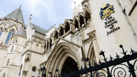 A general view shows a side-on view of the Royal Courts of Justice in London, with a sign on the side of the building showing the coat of arms and a blue cloudy sky in the background.