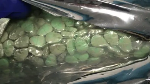 National Crime Agency A clear bag contains lots of green pills in a skull shape. They are sealed in a foil bag