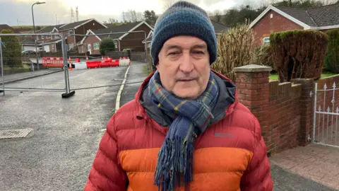 Simon Williams is standing outside the cordoned off road, wearing a red and orange winter coat, tartan scarf and blue and grey striped woolly hat. Metal barriers and a second orange and white safety barrier can be seen behind him.