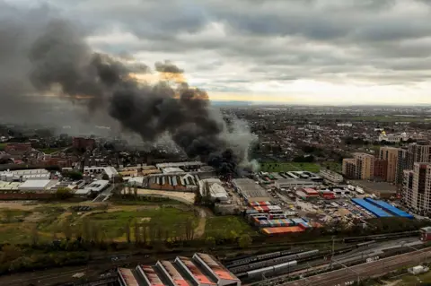 PA Media Smoke rises from a building beyond a railway line. Green scrubland and a car park can be seen either side