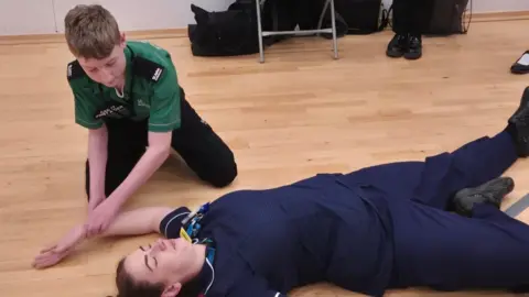 Bedford Giving A young boy, placing someone in the recovery position, wearing a green uniform and black trousers. A person in a blue uniform is lying on the floor, with their eyes closed. 
