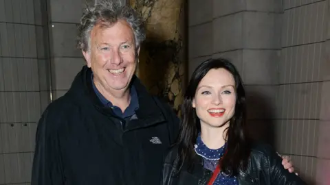 Getty Images A man with wavy greying hair is dressed in a black jacket and blue shirt, and has his arm round a woman with long black hair and wearing a leather jacket.