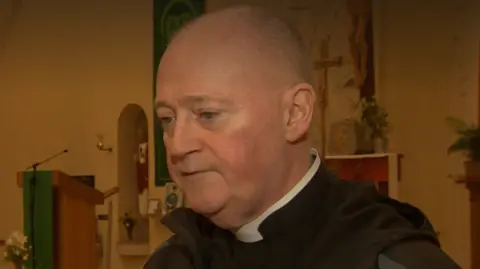 Father Paddy is in Corpus Christi Chapel. He's wearing his black robes and looking away from the camera.