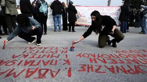 Getty Images Two women write the names of victims of the Tempi crash on the ground outside the courthouse in red paint