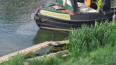Cheney Payne A close up of an end of a house boat on a river. The concrete along the riverbank is falling away into the water. Green grass 