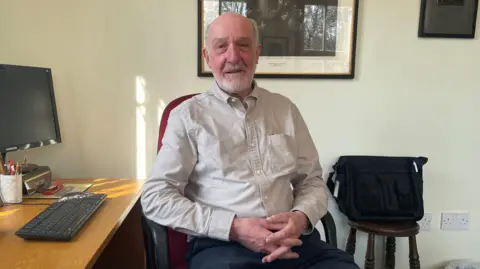 BBC/Jack Hadaway-Weller A man sits on an office chair next to a desk with a computer monitor and keyboard. He is wearing a beige shirt and has his hands folded in his lap. A framed picture hangs on the wall behind him.