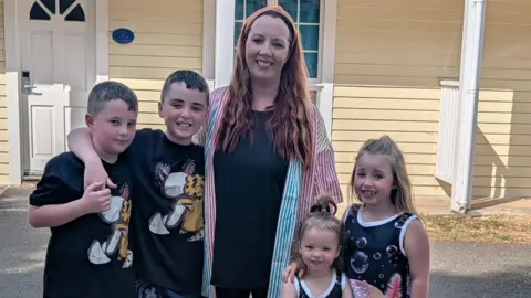 Craig Hinde Trish Hinde stands outside with her children, who are all smiling into the camera. 