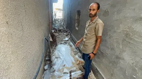 A man stands in an alleyway between two concrete buildings. He has dark hair and a beard and is wearing a short sleeve, light-brown shirt and jeans, holding a mobile phone and is gesturing towards the ground. By his feet is a white plastic sheet covered with some debris and dust, with a corrugated piece of metal leaning against the wall - which constitutes his brother's grave. It is surrounded by a mess of rubble and pieces of metal, the detritus from bombed out buildings nearby.