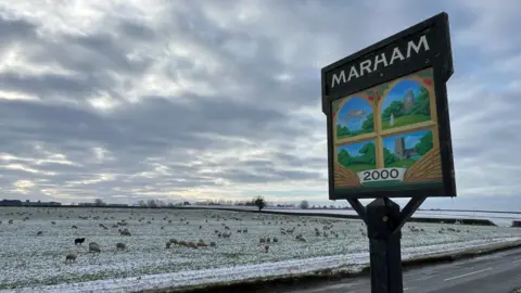 Shaun Whitmore/BBC A village sign says the word Marham on it and the year 2000. It is on a wooden post at the side of a road and there are colourful images of parts of the village on the sign. In the background is a flat, partially show-covered field with lots of sheep grazing