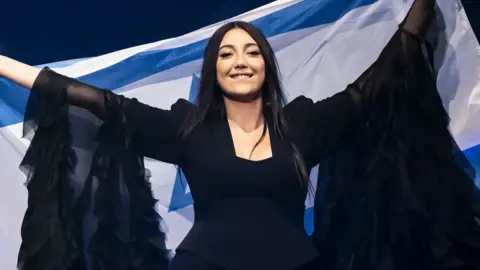 Yuval Raphael smiling and holding up an Israeli flag behind her