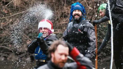 Sam Hepburn A man in a Santa hat, glittery pink wig, life jacket and waterproofs is spraying a watergun at a man with long hair and beard in the foreground. Another man with a blue hat, blue glittery wig, life jacket and waterproofs is laughing. Another man wearing a green wig is using a long wooden oar to paddle. A leafy riverbank can be seen in the background.