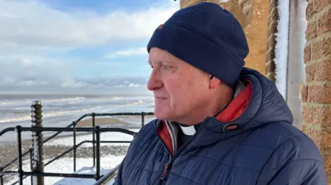 Kevin Shoesmith/BBC A reverend, wearing a black shirt with white dog collar under a navy jacket, looks out to sea. He has a sombre, concerned expression.