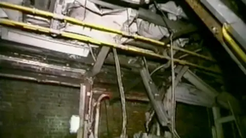 PA Media Wreckage hangs from the ceiling of a rube carriage. The yellow handrails are in tact across the ceiling, but debris is falling around it. In the background are the brick walls of the London Underground.