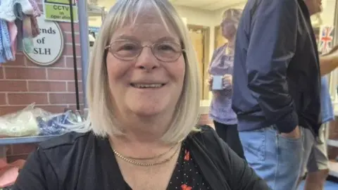 Christine is smiling and had blond hair in a bob which stops just below her chin. She is wearing a gold necklace and a black top which has little red and white spots on it. Behind her a few people standing an talking.
