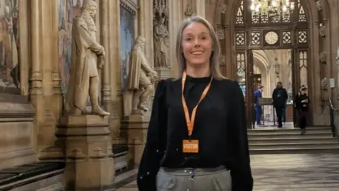 A woman with fair hair, wearing an orange "visitor" lanyard over a black top, stands in an ornate hallway in Parliament with statues along the walls.