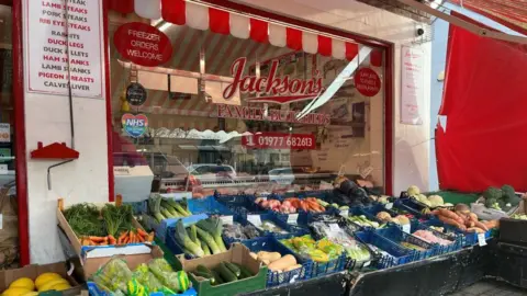 BBC/Julia Lewis A red and white shop front which says "Jackson's family butchers".
There's baskets of vegetables laid out in baskets in front of the window including leeks, carrots, sweet potato, courgettes and broccoli.