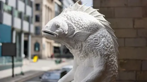 A close-up of the sculpture Skalli which is white in colour and located on a plinth at the side of Liverpool Parish Church. It is in an elevated position with a road with traffic passing buy below.