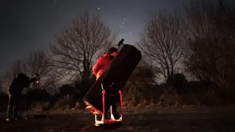 Simon Holbeche Ben Luger observing at Wellow village with Bath Astronomers. He is looking through a professional grade telescope and is standing on the right side of it, wearing a black beanie hat and thick clothing. The trees are silhouetted against a starlit sky.
