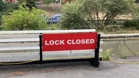 Emma Howgego/BBC A big red sign with the words "lock closed" written in white capital letters. White wooden fencing is behind the sign and a brick wall. Beyond that are some trees, with a river and houseboat visible. 