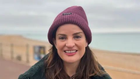 George Carden/BBC A woman with long brown hair looking and smiling at the camera. She is wearing a fark fluffy coat and a purple beanie. The beach and sea is behind her.