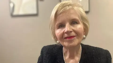 Donna Ockenden looking at the camera. She has short blonde hair and is wearing a black top with a string of pearls.