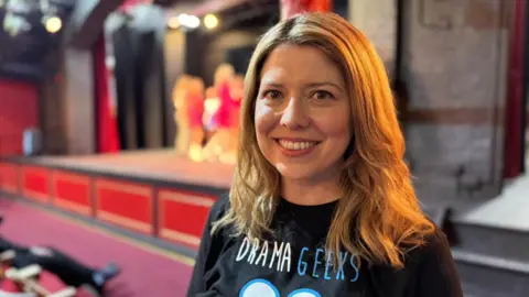 Charlotte Reid smiles at the camera. She has shoulder length blond hair and is wearing a black t-shirt with "Drama Geeks" written on it in white and blue letters. Behind her is a stage with several young people on it.